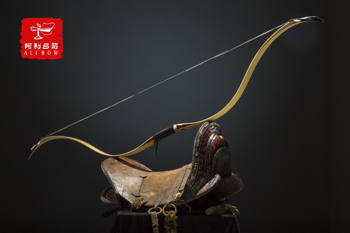 A traditional laminated bow, strung, resting across an antique leather saddle against a dark backdrop.